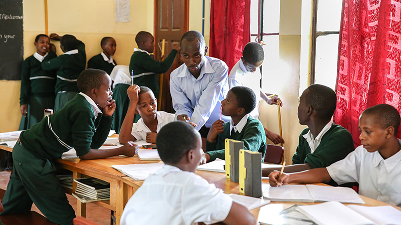 A group of Tanzanian students gather around a table. Some listen to their mentor who is perched on the end of the table talking, while others work on their tablets.