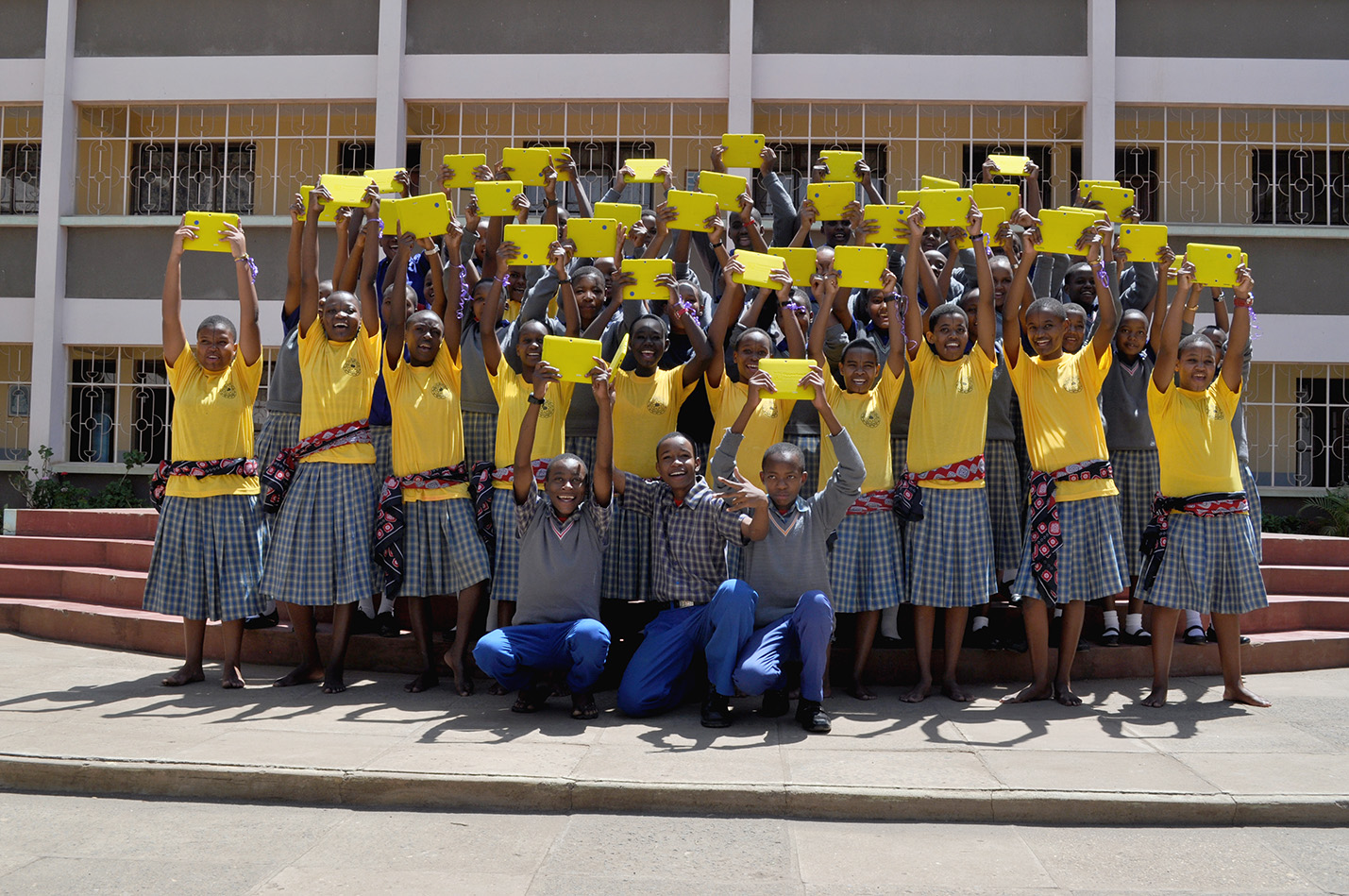 A class of students smiles while proudly holding their bright yellow Opportunity Tablets high.