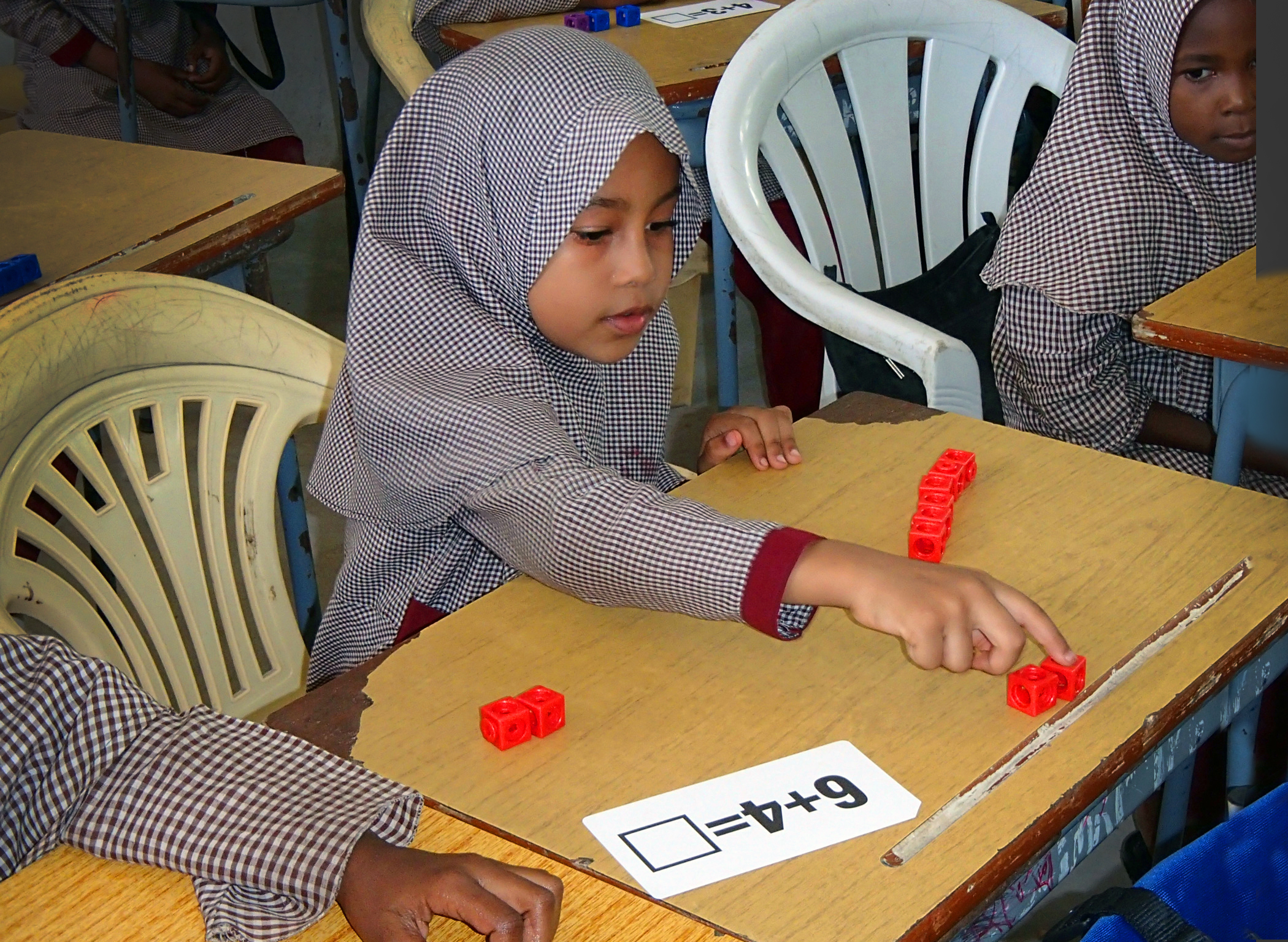 A Primary Program student counts with blocks. In front of her is a card that reads "6+4=[]."