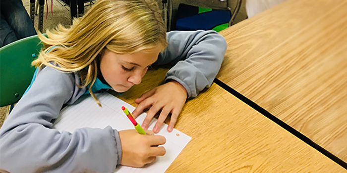 A student writes a pen-pal letter to a Sister School student at her desk with a colorful pencil and lined paper.