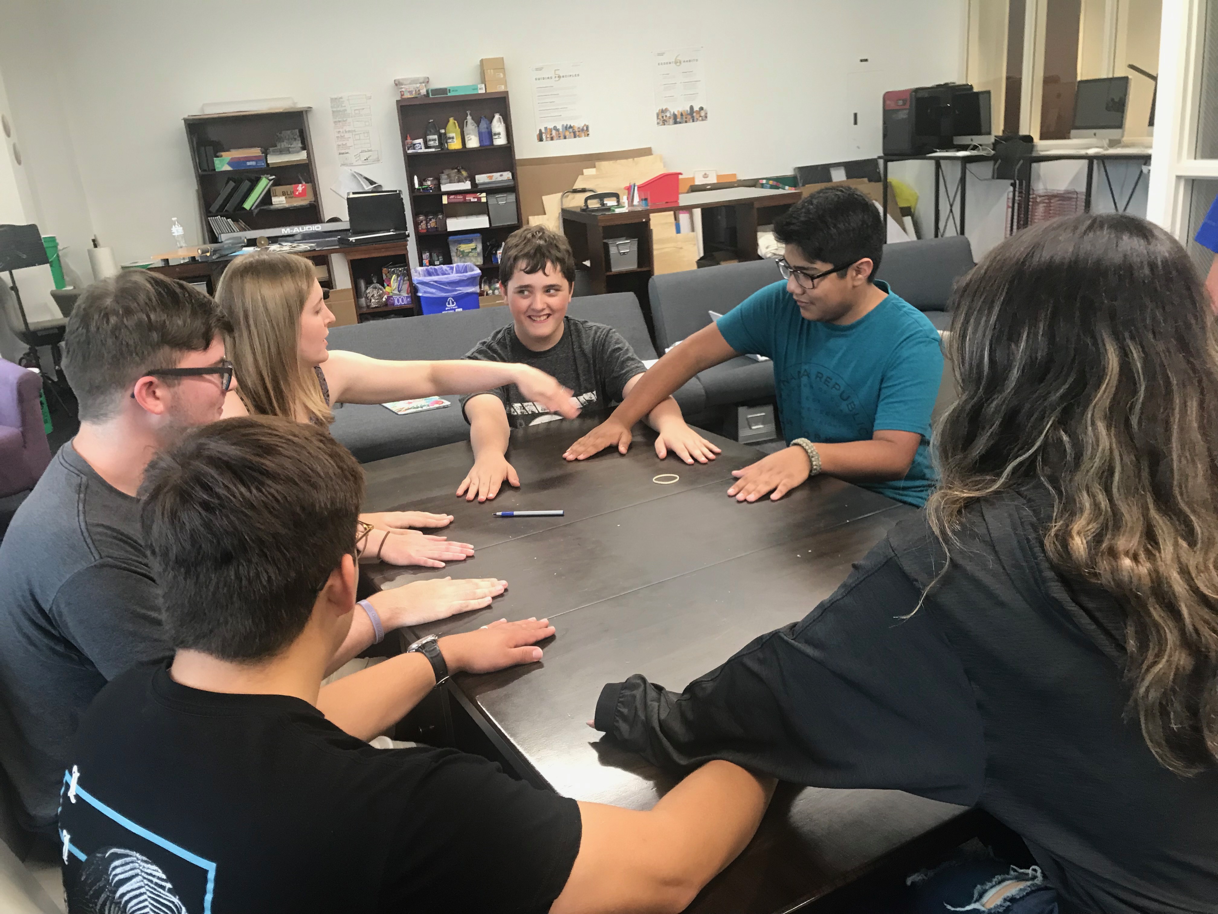 Students gather around a table and play a game where they overlap their arms with one another.