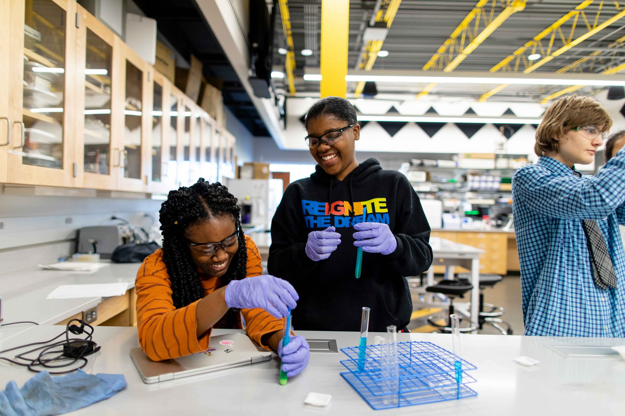 In the science lab, two students examine test tubes while smiling and laughing. 