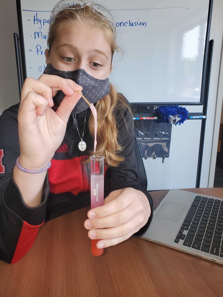 A female student wears a mask and holds a test tube, excitedly and carefully pulling out a pink substance from it.