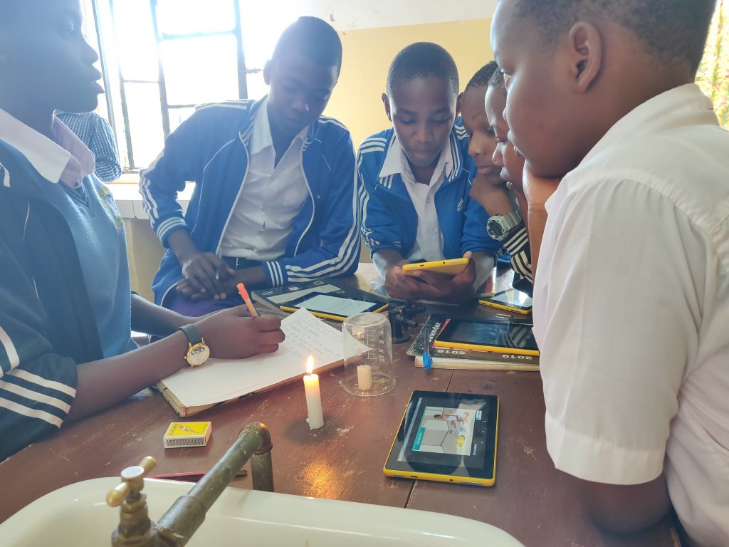Students gather around a work table, with touch-screen tablets and two candles, one lit and one extinguished under a beaker, on the table in front of them.