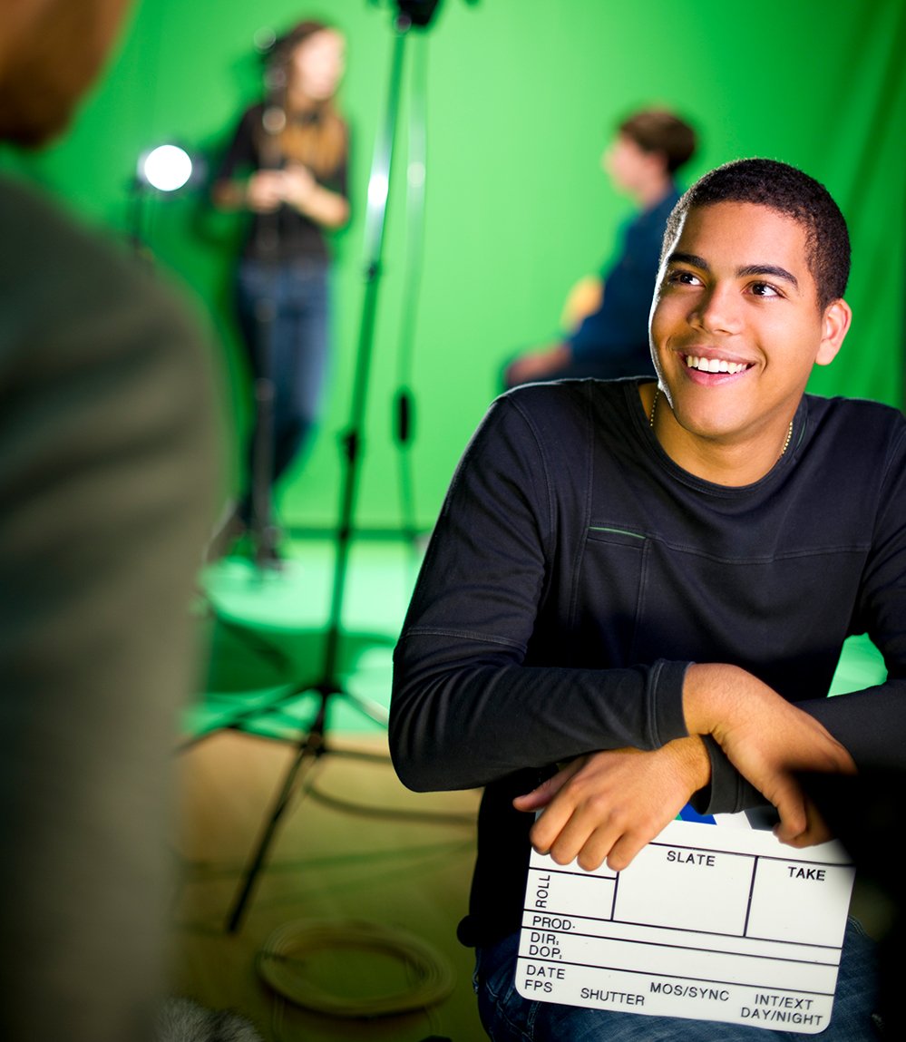 A student smiles while looking up at an internship colleague on a film set, with a green screen and lighting behind them.