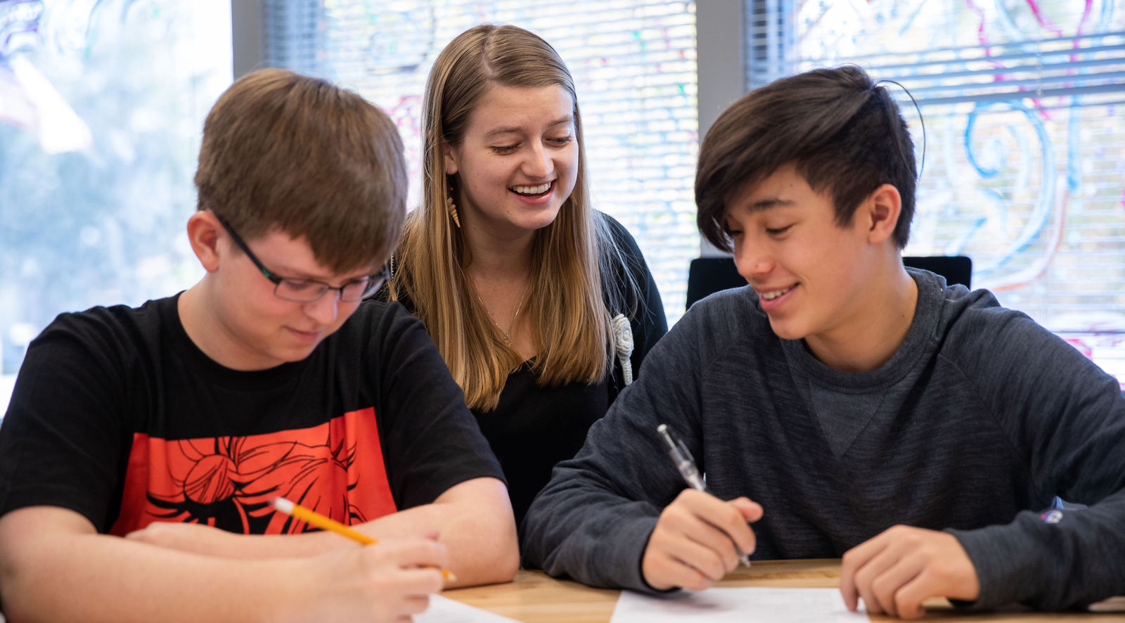 A mentor smiles with two students working at a table.
