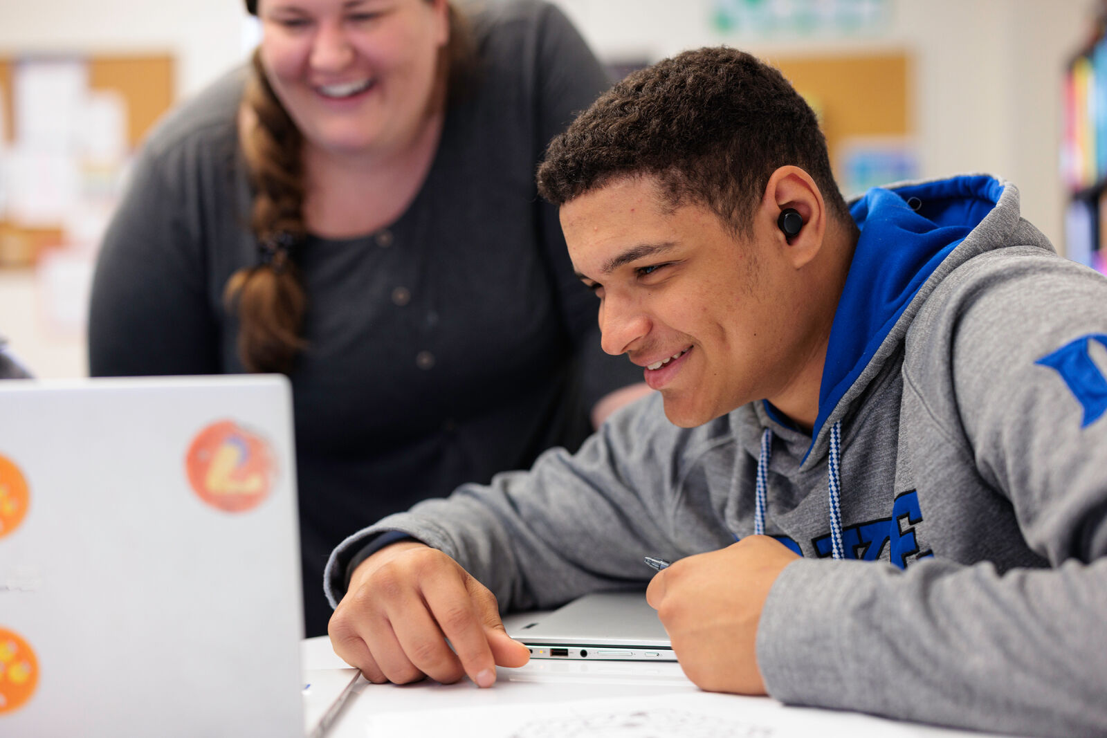 A student smiles while looking at a laptop with his teacher.