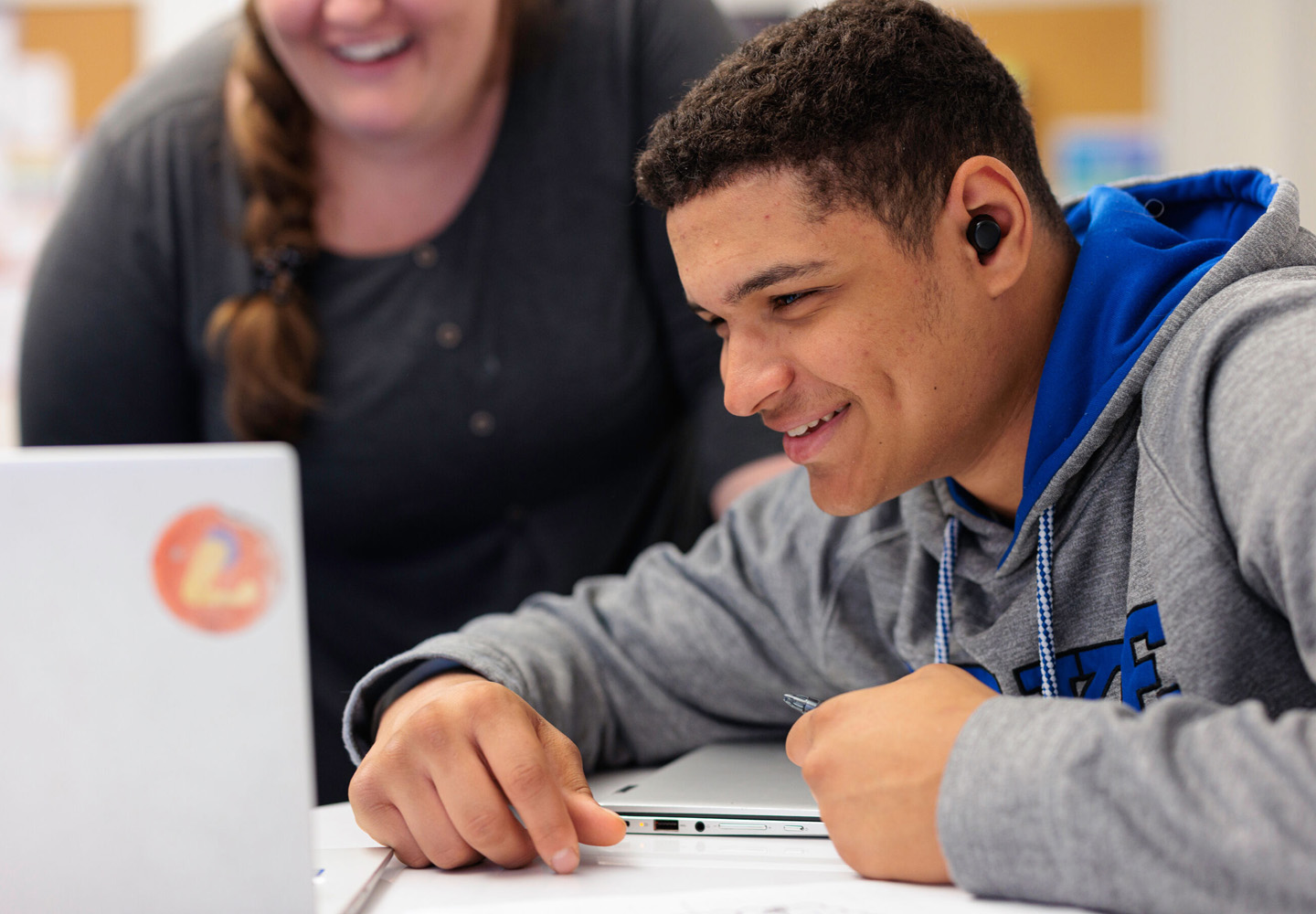 A student smiles as he looks at a laptop in front of him, with a teacher smiling in the background